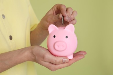 Photo of Woman putting coin into pink piggy bank on olive background, closeup