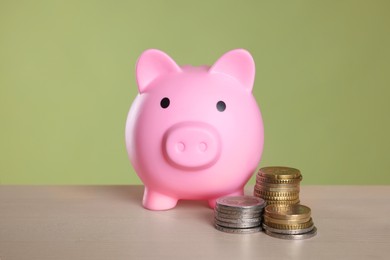 Pink piggy bank and coins on wooden table against olive background