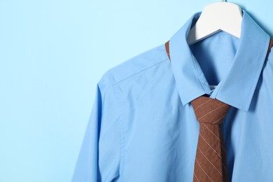 Photo of Hanger with shirt and brown necktie on light blue background, closeup. Space for text