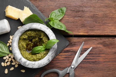 Photo of Tasty pesto sauce in mortar, basil, pine nuts, cheese and scissors on wooden table, top view