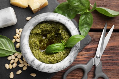 Photo of Tasty pesto sauce in mortar, basil, pine nuts, cheese and scissors on wooden table, top view