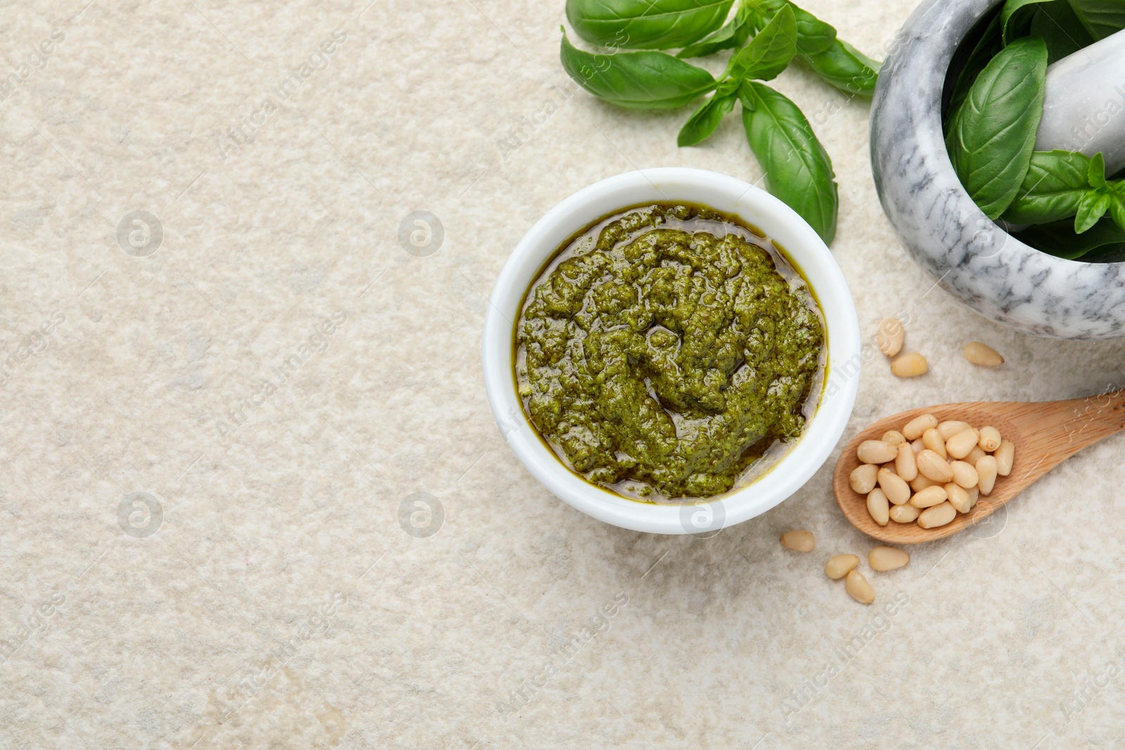 Photo of Tasty pesto sauce in bowl, basil, pine nuts and spoon on light grey table, flat lay. Space for text