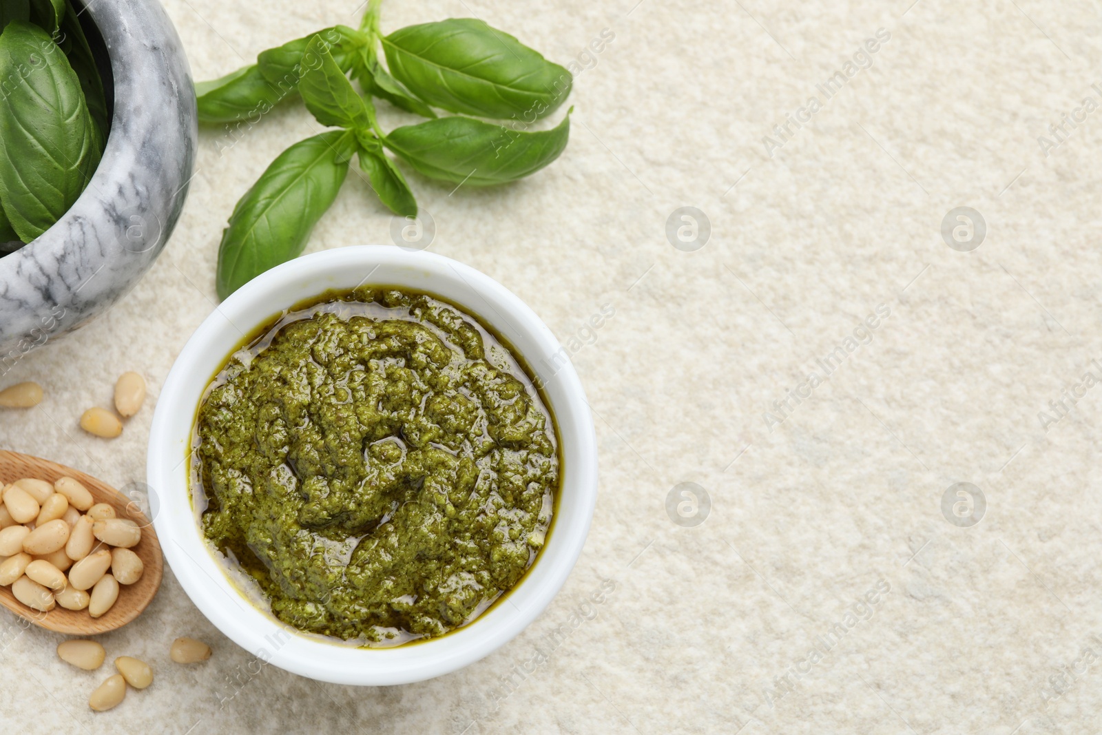 Photo of Tasty pesto sauce in bowl, basil, pine nuts and spoon on light grey table, flat lay. Space for text