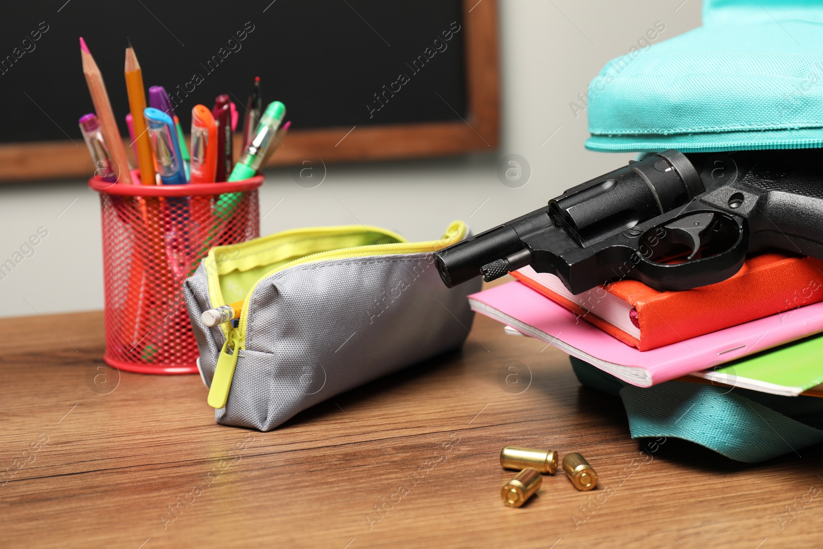 Photo of Gun, bullets and school stationery on wooden table near blackboard indoors, closeup