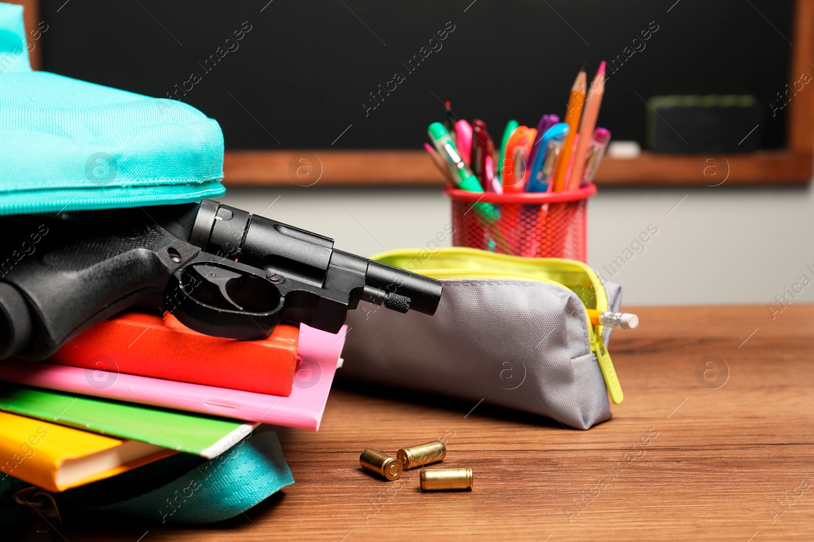 Photo of Gun, bullets and school stationery on wooden table near blackboard indoors, closeup