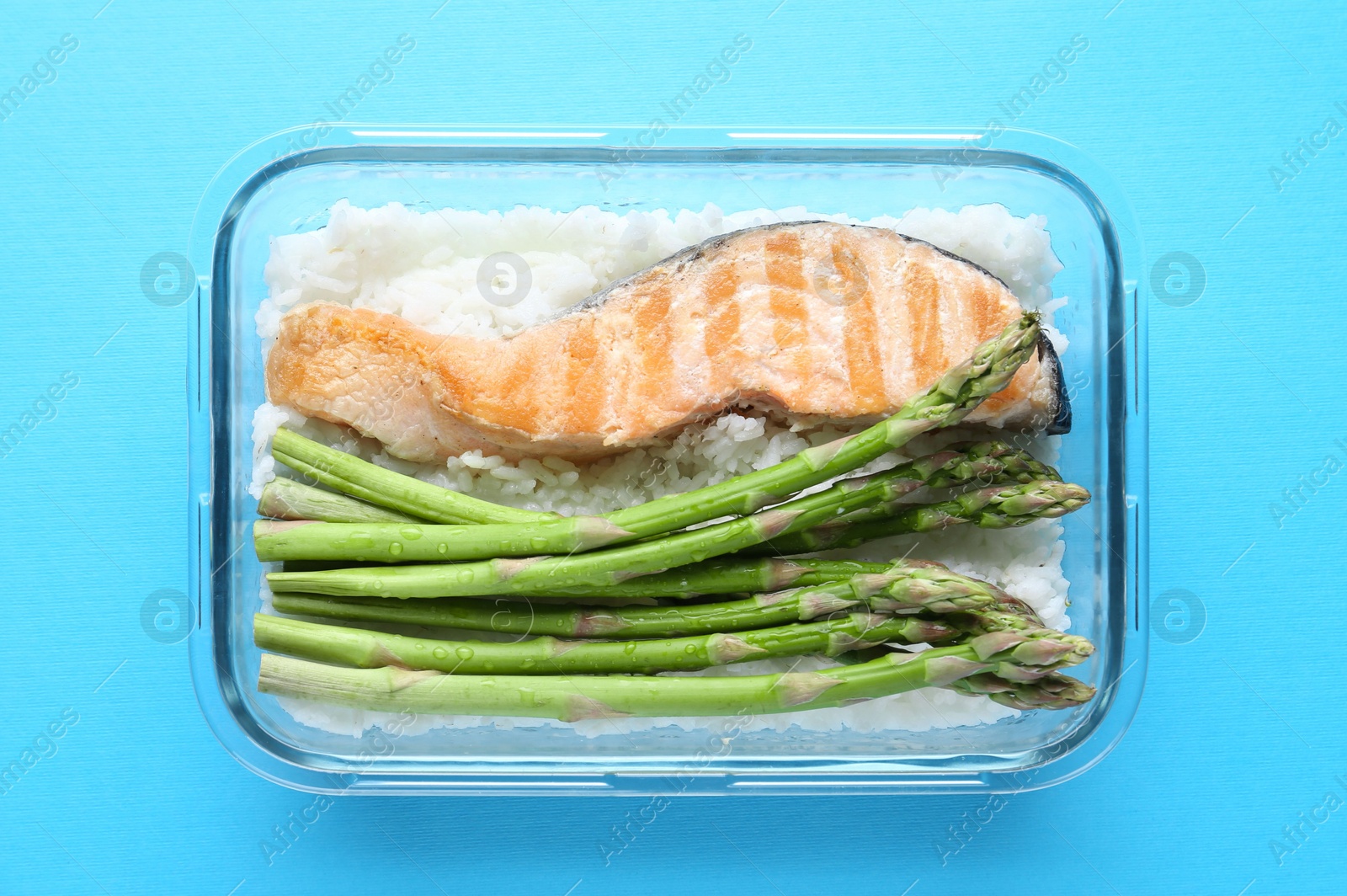 Photo of Healthy meal. Fresh asparagus, salmon and rice in glass container on light blue background, top view