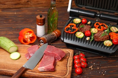 Electric grill with meat, spices, knife and vegetables on wooden table