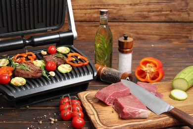Electric grill with meat, spices, knife and vegetables on wooden table
