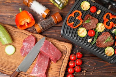 Photo of Electric grill with meat, spices, knife and vegetables on wooden table, flat lay