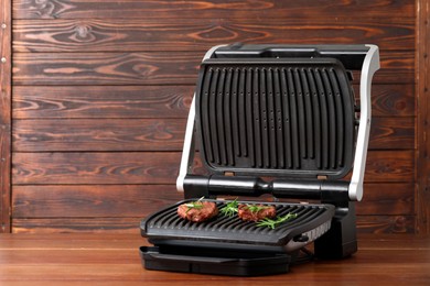 Photo of Electric grill with tasty meat steaks and rosemary on wooden table. Space for text