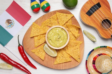 Delicious guacamole with nachos chips, Mexican flag, sombrero hat, ukulele and maracas on white wooden table, flat lay