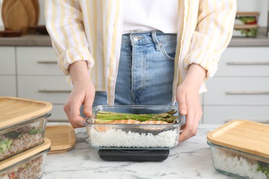 Photo of Healthy meal. Woman weighing glass container with food on kitchen scale at white marble table, closeup