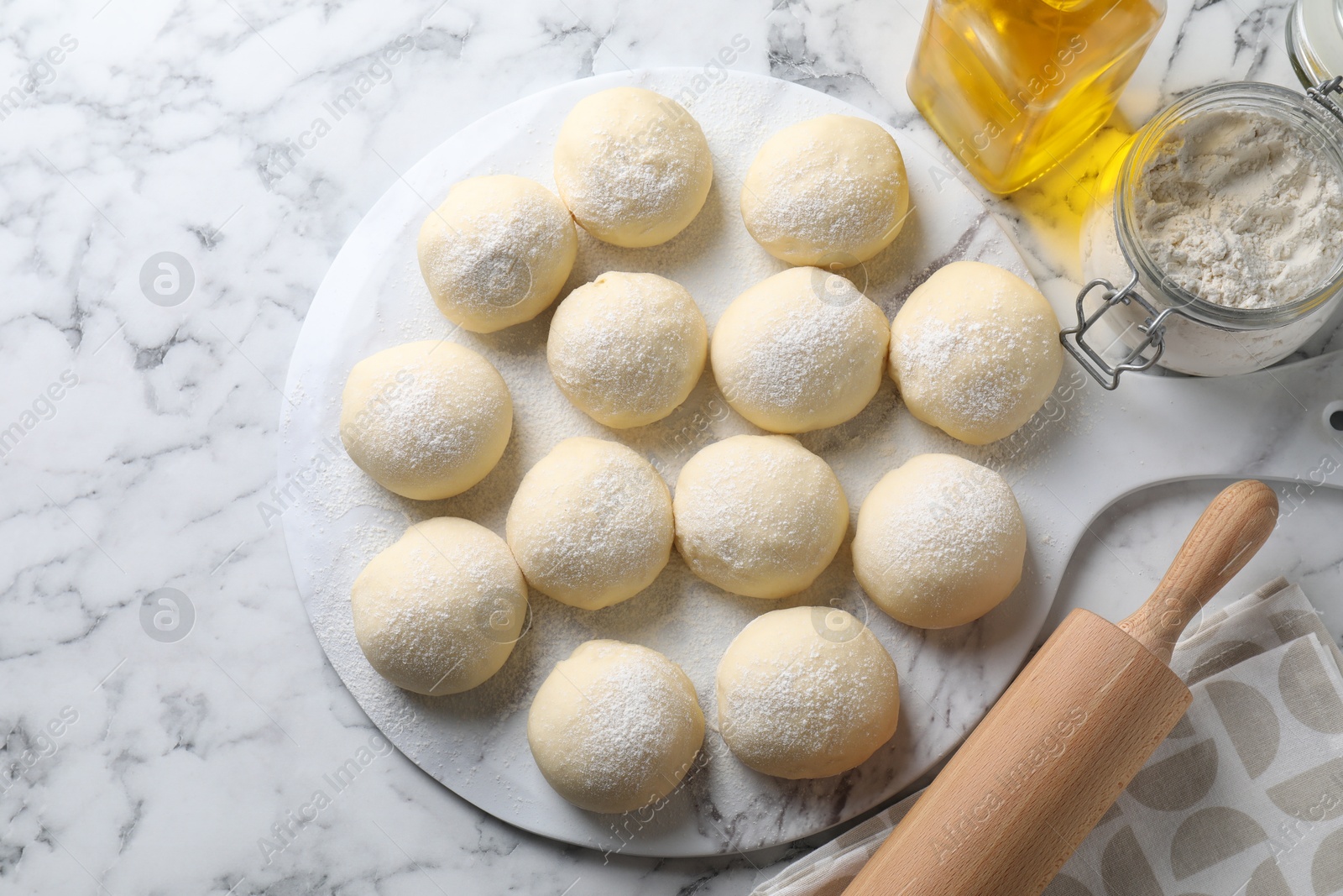 Photo of Raw dough balls, flour, rolling pin and oil on white marble table, top view