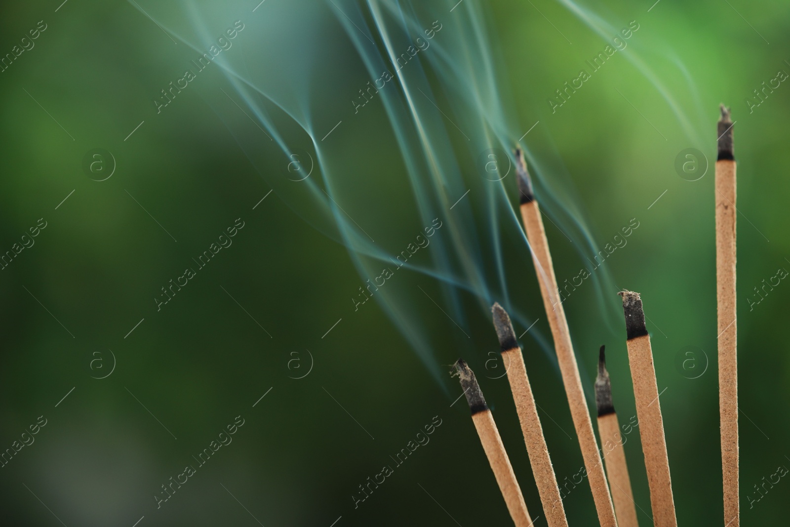 Photo of Incense sticks smoldering on green blurred background, space for text