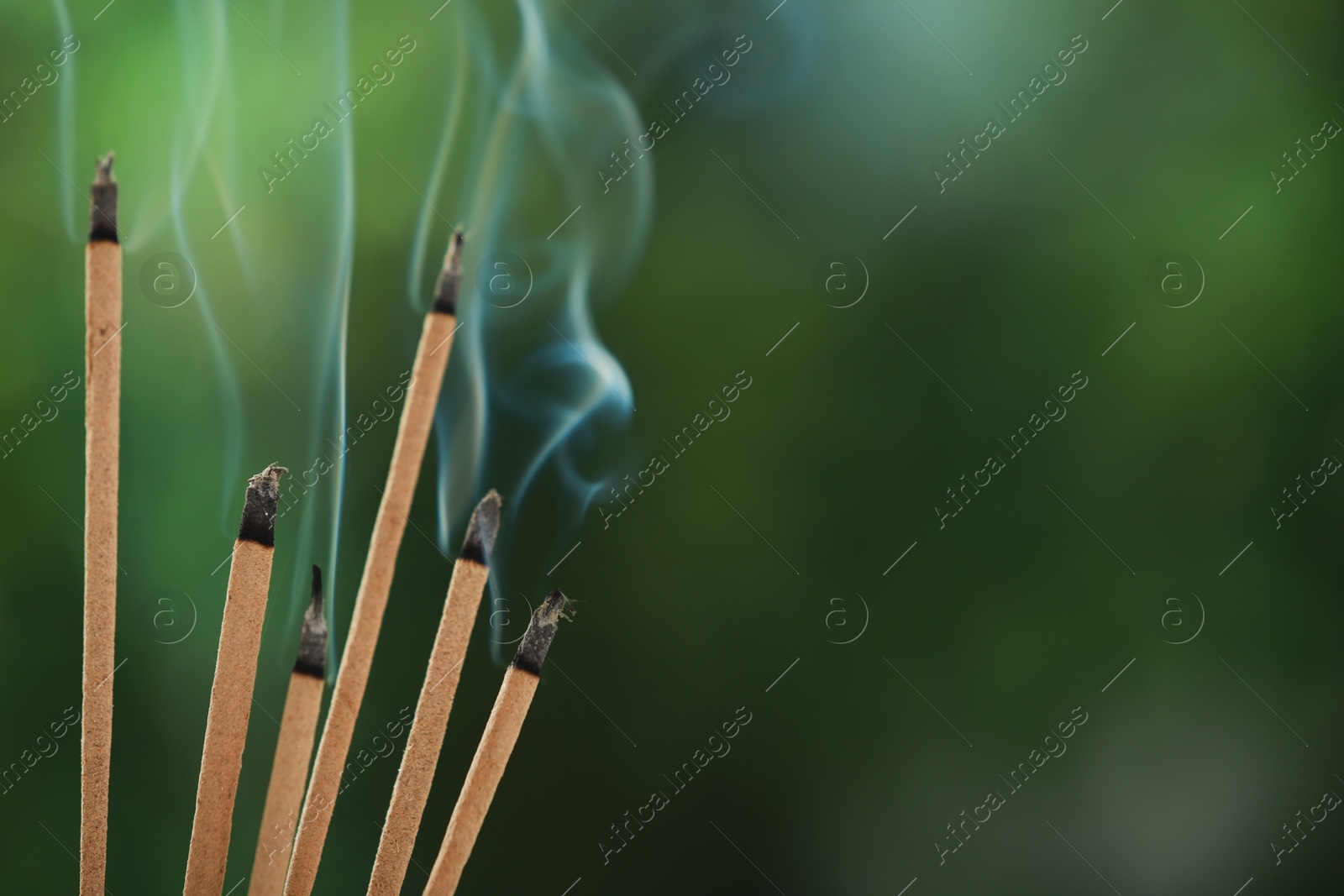 Photo of Incense sticks smoldering on green blurred background, space for text