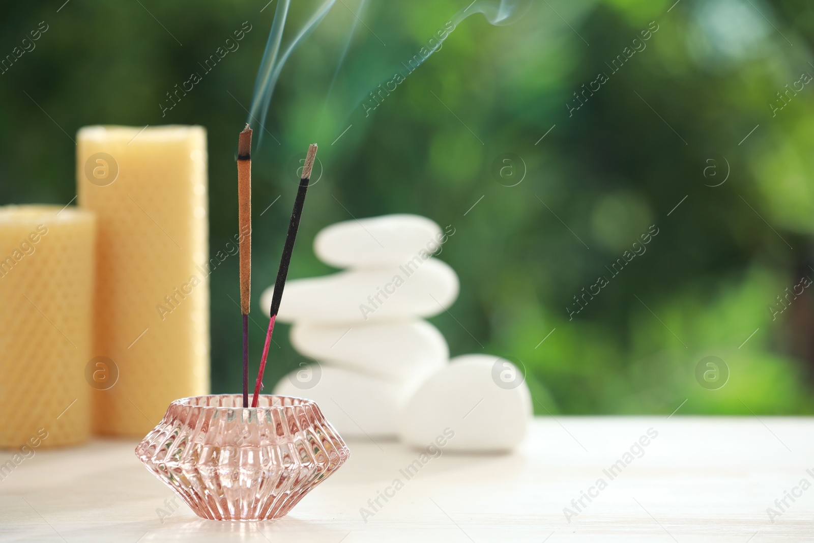 Photo of Incense sticks smoldering in holder near stones and candles on wooden table outdoors, space for text