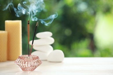 Incense sticks smoldering in holder near stones and candles on wooden table outdoors, space for text