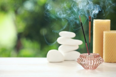 Photo of Incense sticks smoldering in holder near stones and candles on wooden table outdoors, space for text