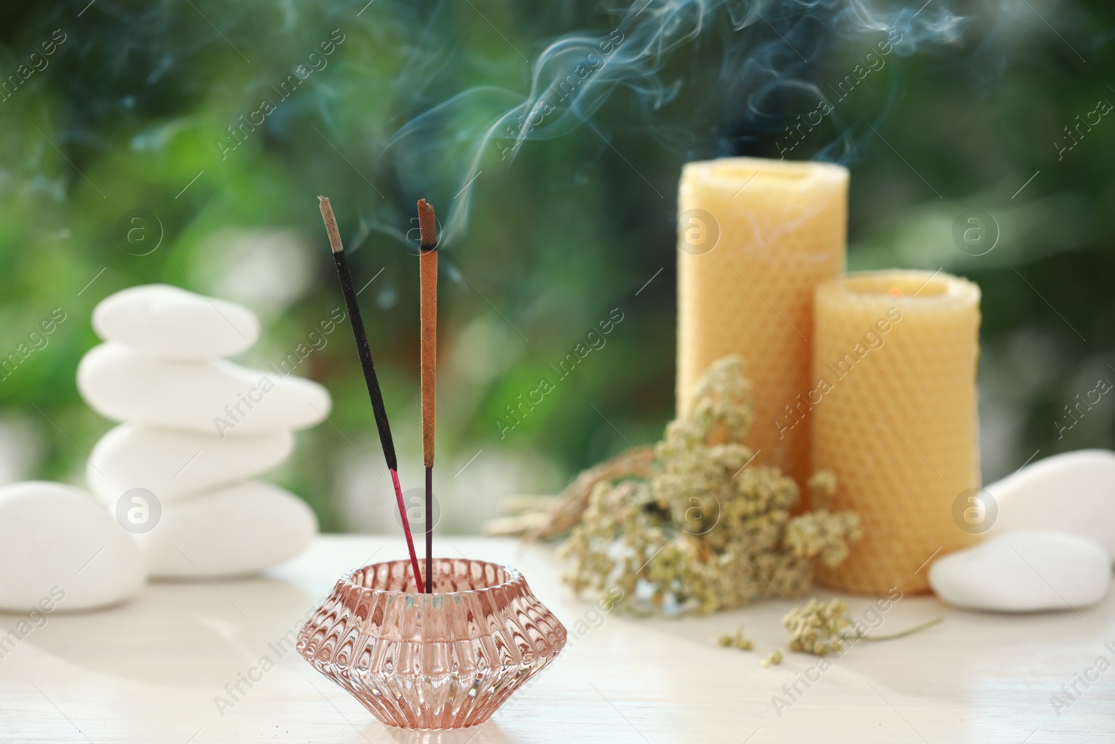 Photo of Incense sticks smoldering in holder near stones, candles and dry flowers on wooden table outdoors