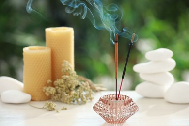 Photo of Incense sticks smoldering in holder near stones, candles and dry flowers on wooden table outdoors