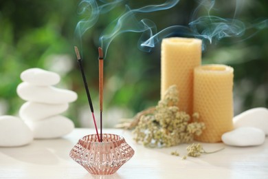 Incense sticks smoldering in holder near stones, candles and dry flowers on wooden table outdoors