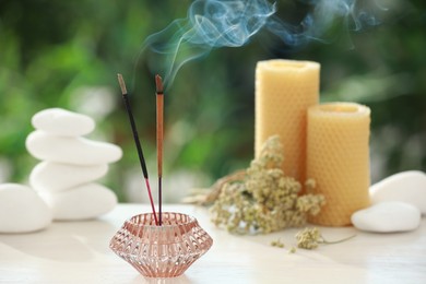 Incense sticks smoldering in holder near stones, candles and dry flowers on wooden table outdoors