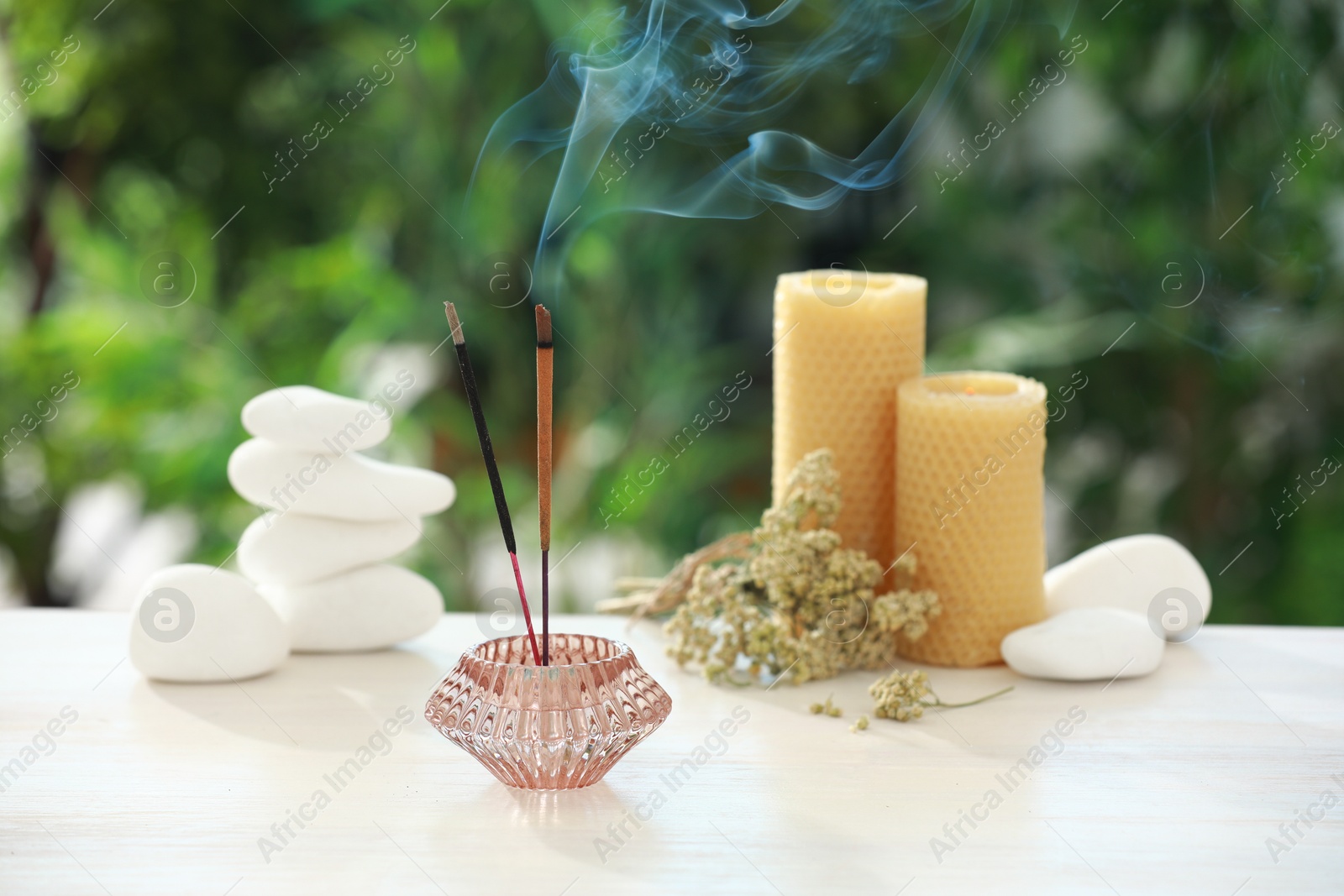 Photo of Incense sticks smoldering in holder near stones, candles and dry flowers on wooden table outdoors
