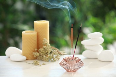 Incense sticks smoldering in holder near stones, candles and dry flowers on wooden table outdoors
