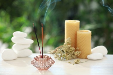 Photo of Incense sticks smoldering in holder near stones, candles and dry flowers on wooden table outdoors