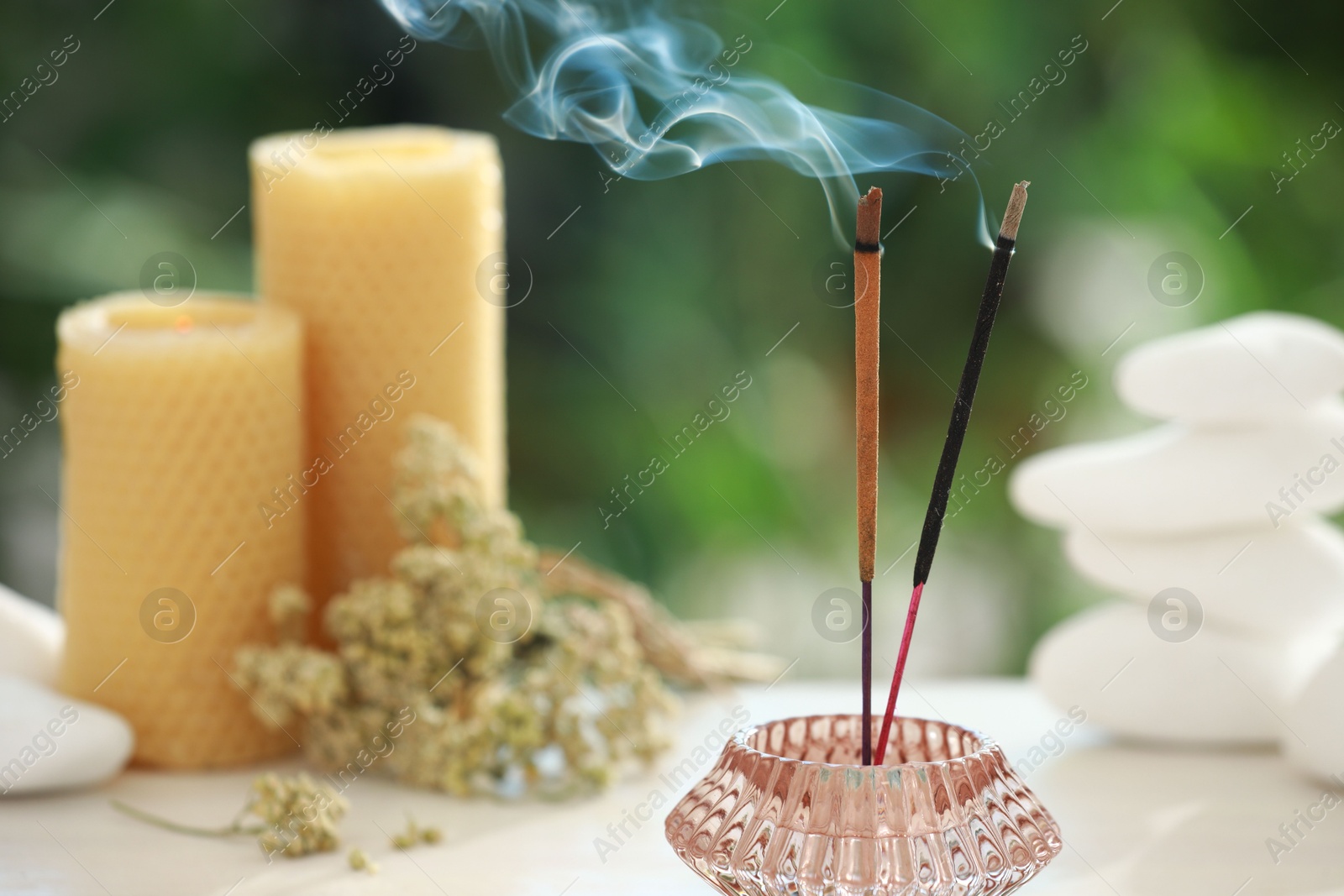 Photo of Incense sticks smoldering in holder near stones, candles and dry flowers on table outdoors