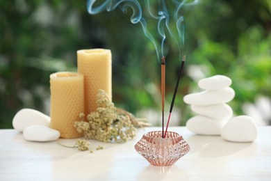 Incense sticks smoldering in holder near stones, candles and dry flowers on wooden table outdoors