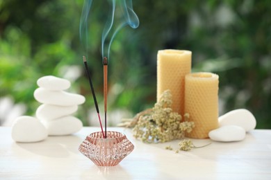 Incense sticks smoldering in holder near stones, candles and dry flowers on wooden table outdoors