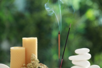 Photo of Incense sticks smoldering near stones, candles and dry flowers against green blurred background