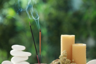 Photo of Incense sticks smoldering near stones, candles and dry flowers against green blurred background