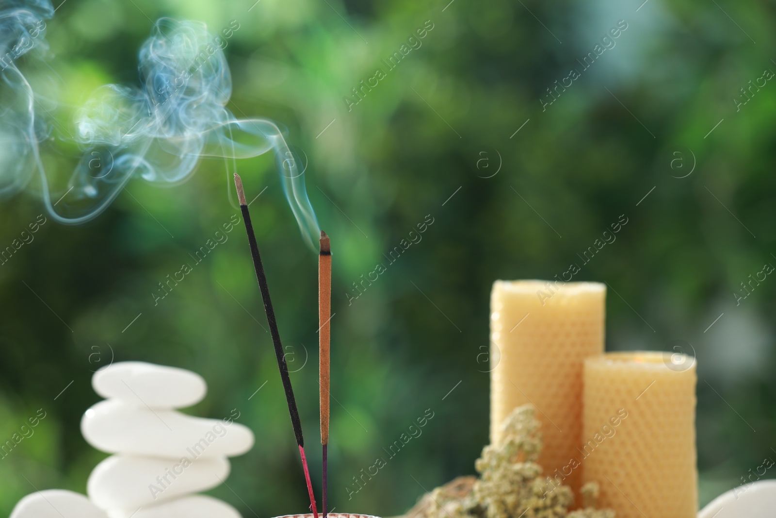 Photo of Incense sticks smoldering near stones, candles and dry flowers against green blurred background