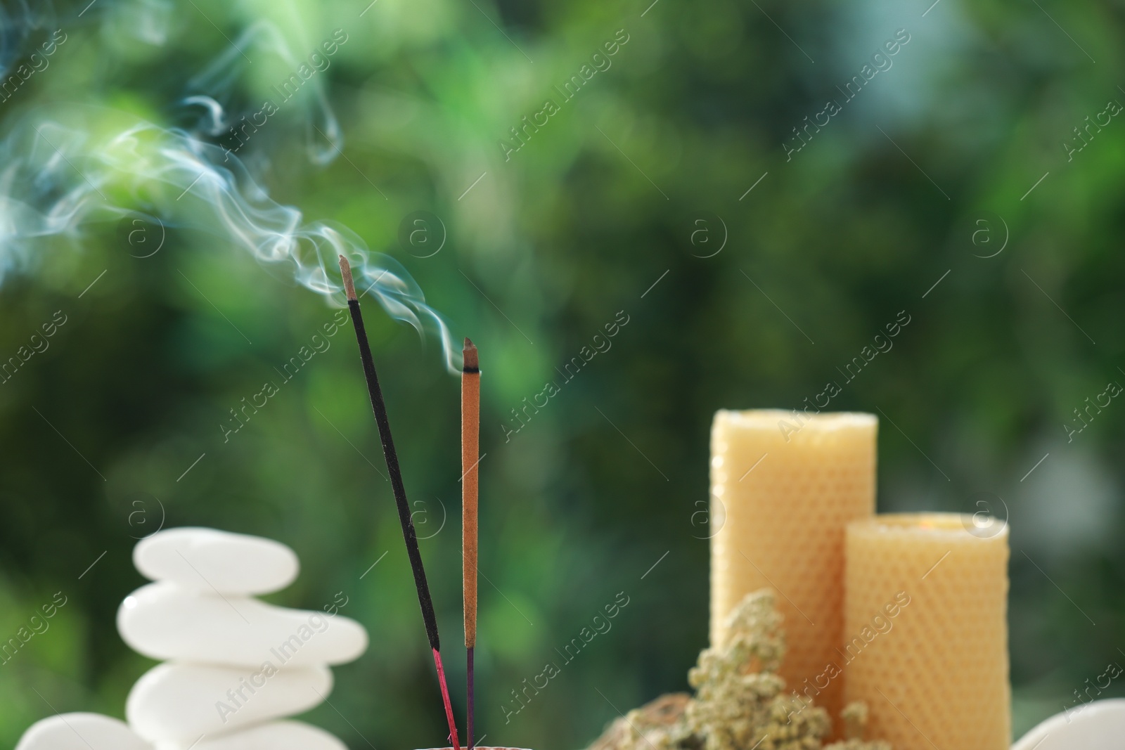 Photo of Incense sticks smoldering near stones, candles and dry flowers against green blurred background