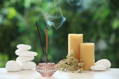 Incense sticks smoldering in holder near stones, candles and dry flowers on wooden table outdoors