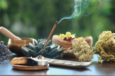 Photo of Incense stick smoldering in holder, candles and dry flowers on wooden table outdoors