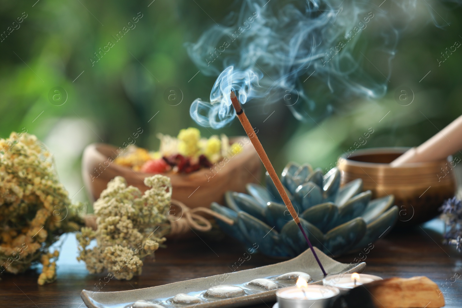 Photo of Incense stick smoldering in holder, candles and dry flowers on wooden table outdoors