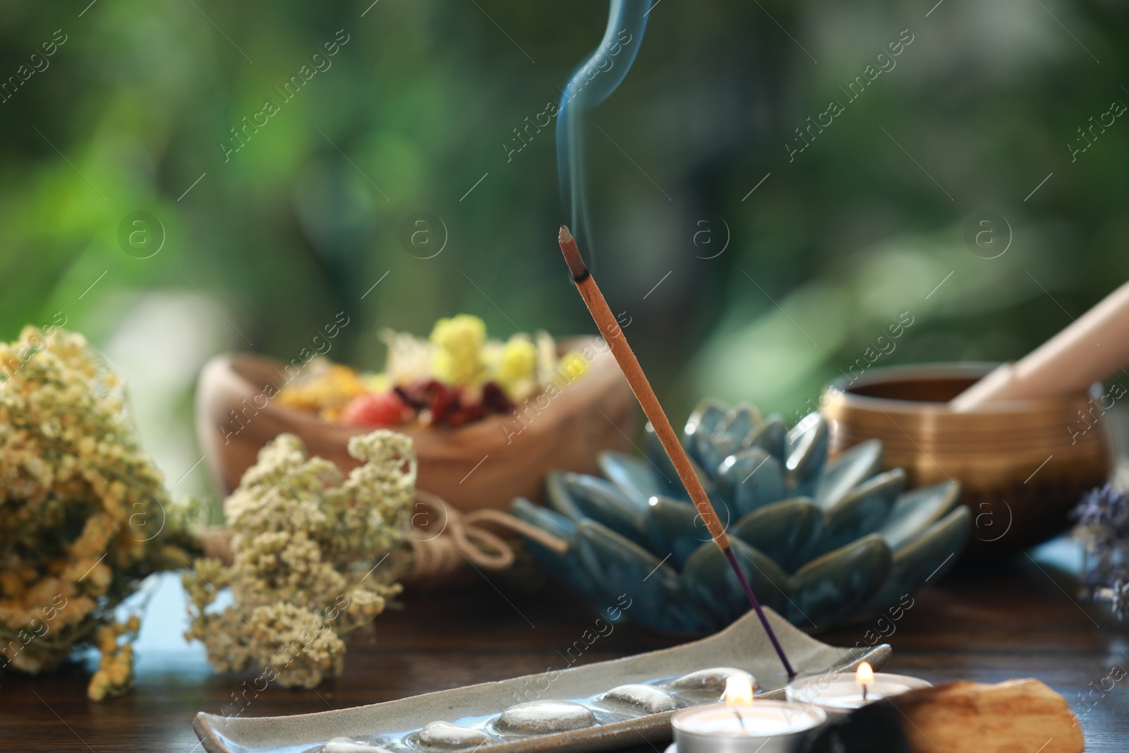 Photo of Incense stick smoldering in holder, candles and dry flowers on wooden table outdoors