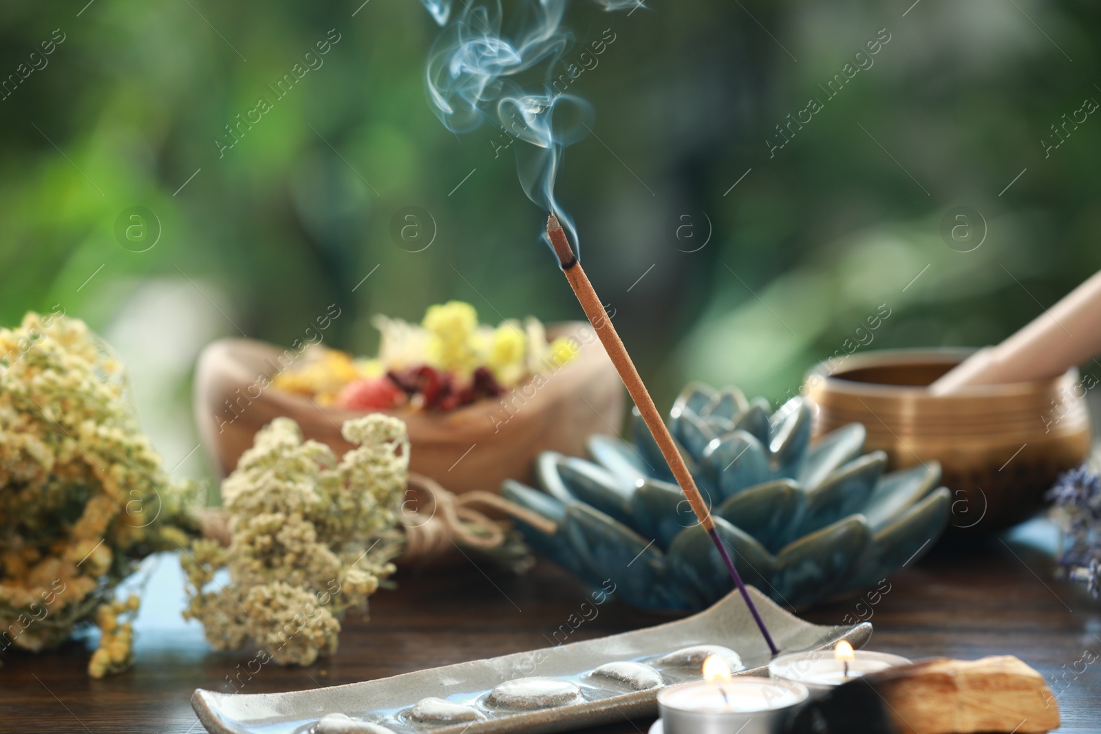 Photo of Incense stick smoldering in holder, candles and dry flowers on wooden table outdoors