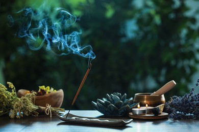 Photo of Incense stick smoldering in holder, burning candles, dry flowers and Tibetan singing bowl on wooden table outdoors