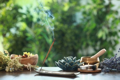 Incense stick smoldering in holder, burning candles, dry flowers and Tibetan singing bowl on wooden table outdoors