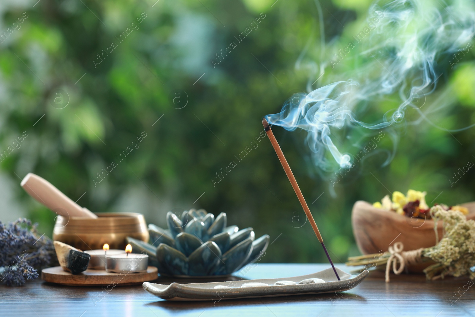 Photo of Incense stick smoldering in holder, burning candles, dry flowers and Tibetan singing bowl on wooden table outdoors