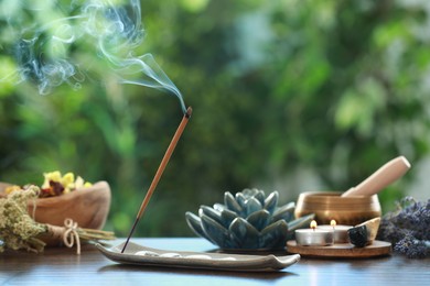 Photo of Incense stick smoldering in holder, burning candles, dry flowers and Tibetan singing bowl on wooden table outdoors