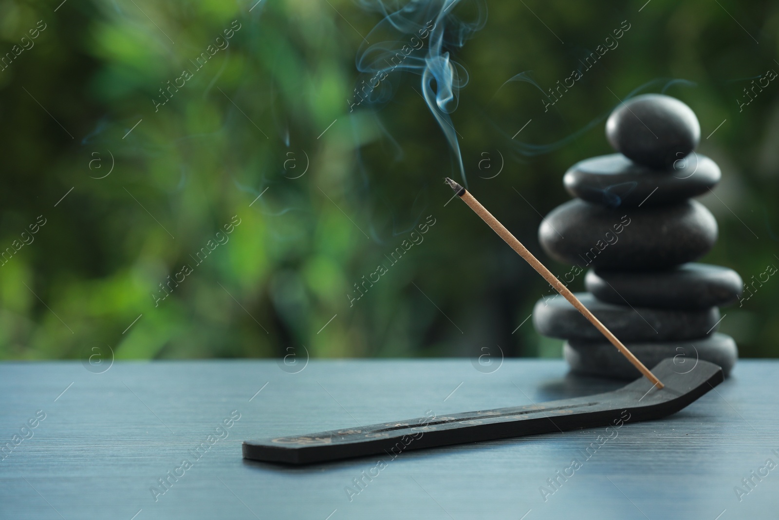 Photo of Incense stick smoldering in holder and spa stones on wooden table outdoors, space for text. Om ligature