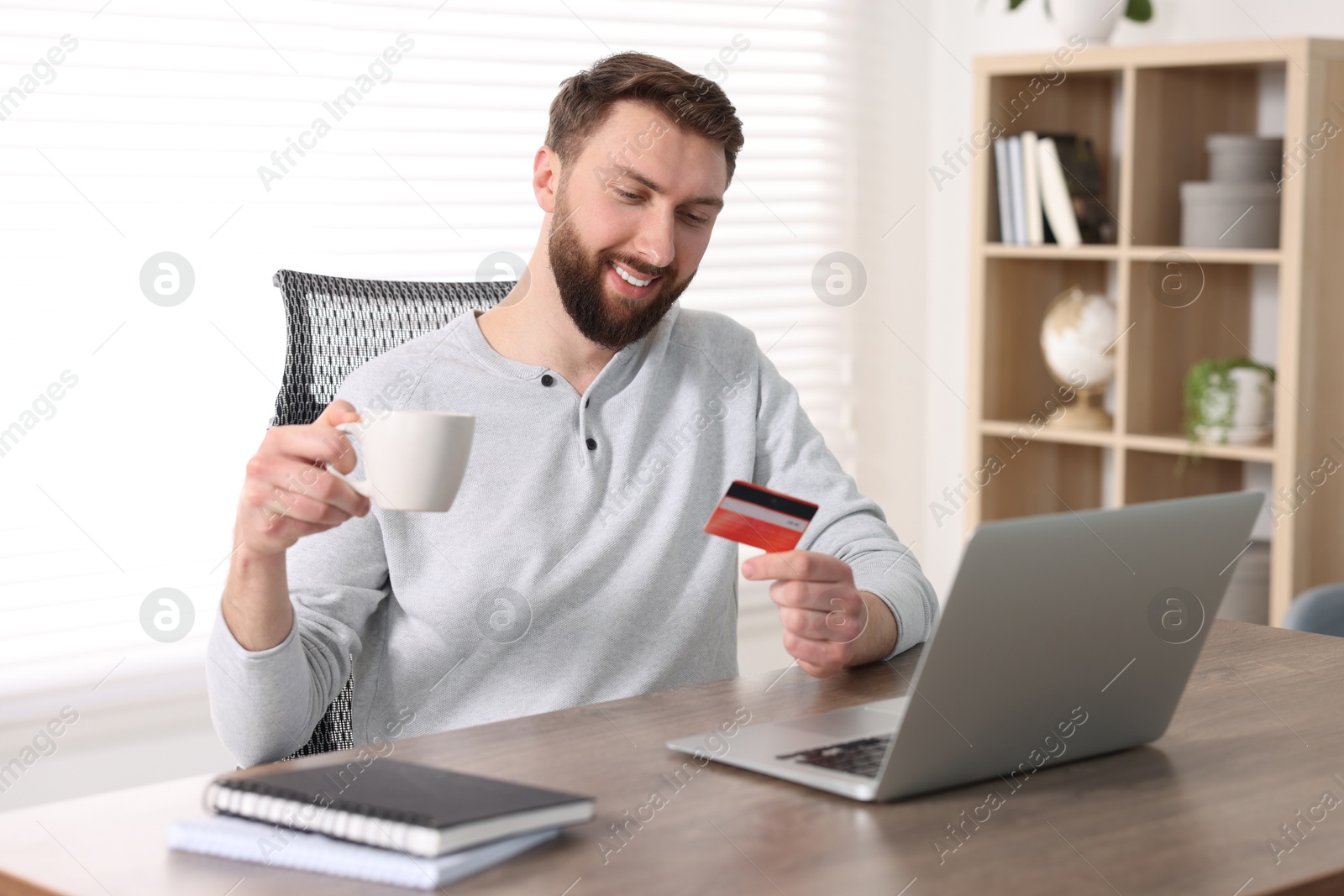 Photo of Online banking. Happy young man with credit card and laptop paying purchase at table indoors