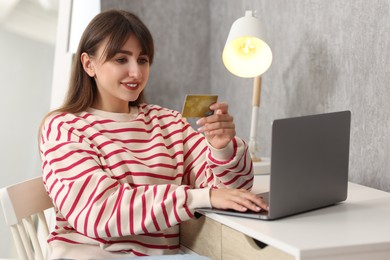 Photo of Online banking. Smiling woman with credit card and laptop paying purchase at table indoors
