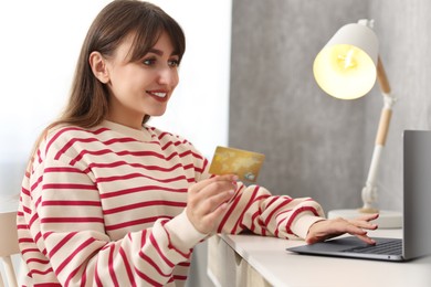 Photo of Online banking. Smiling woman with credit card and laptop paying purchase at table indoors