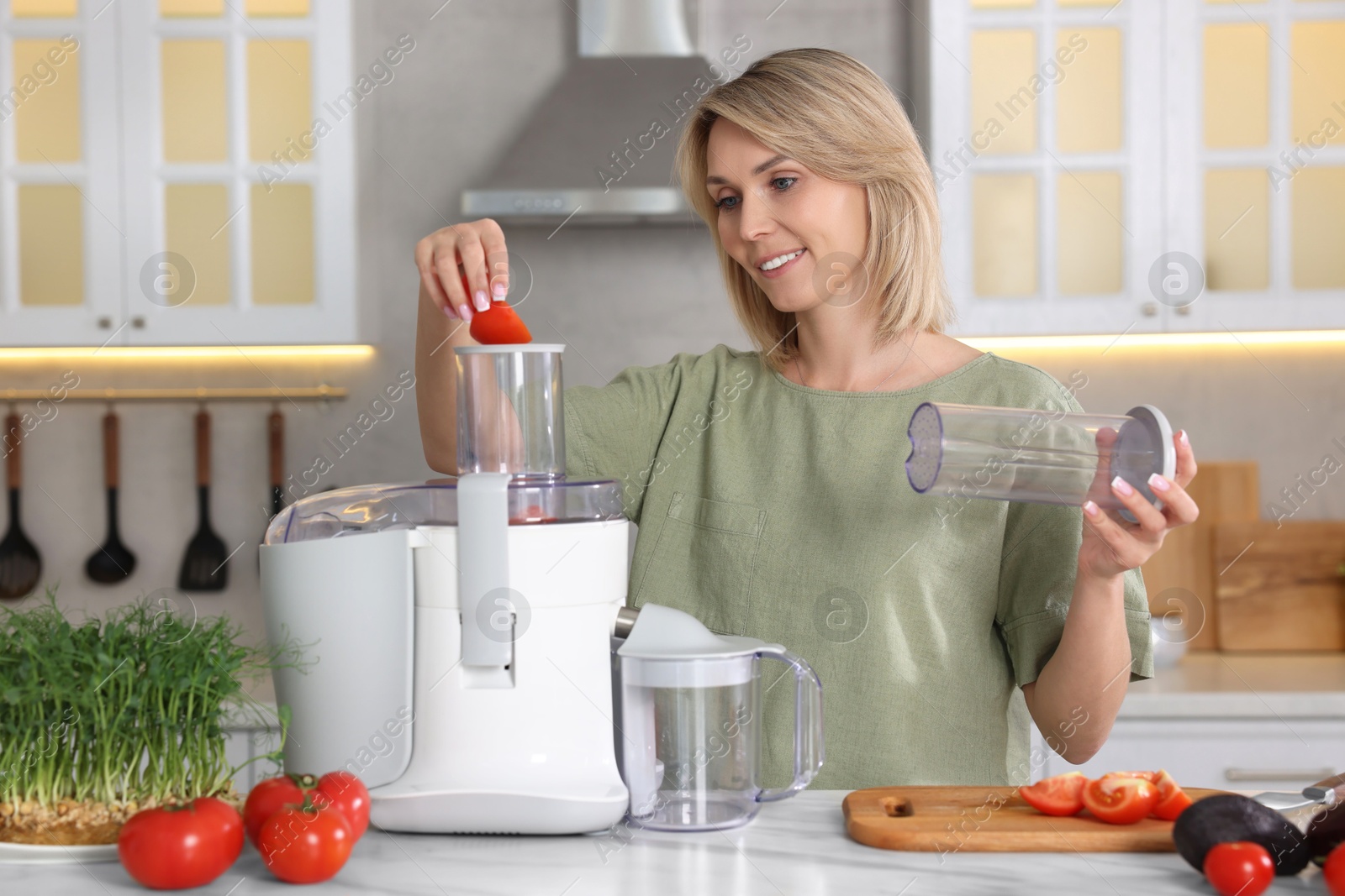 Photo of Smiling woman putting fresh tomato into juicer at white marble table in kitchen